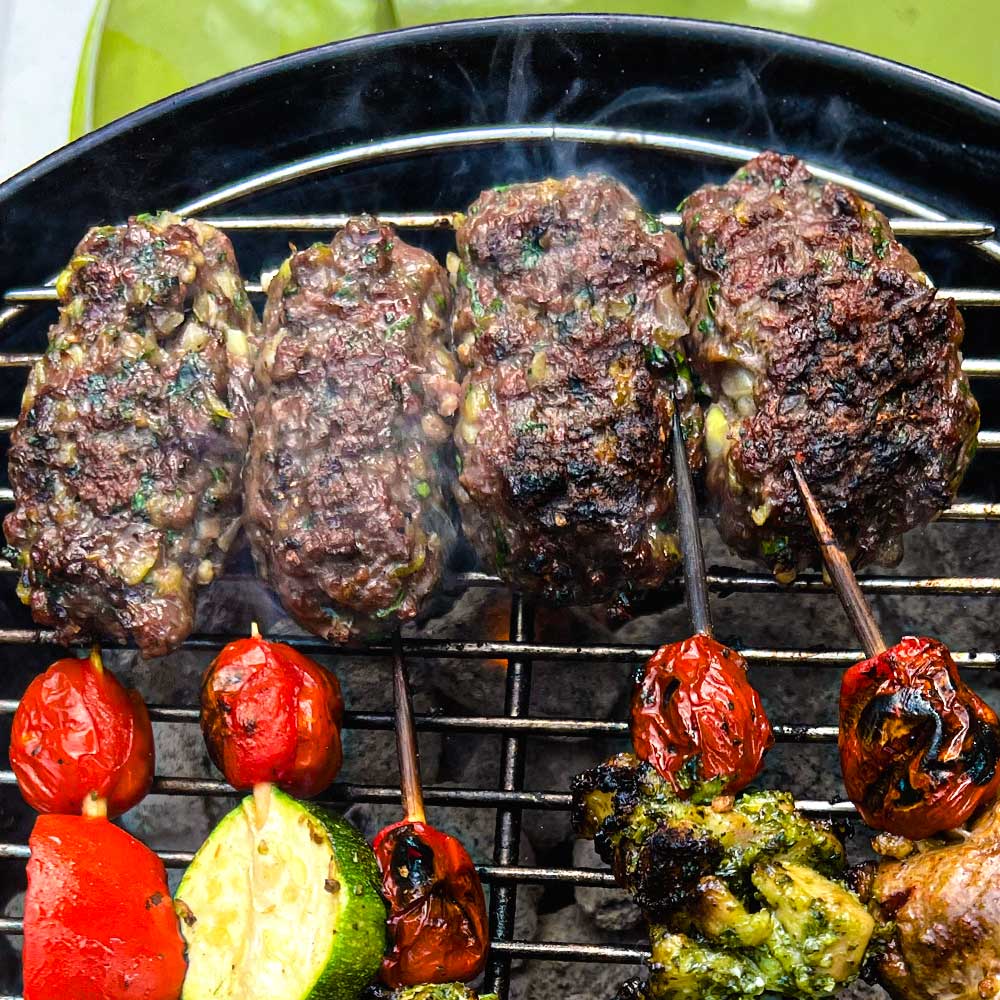 Hot Charcoal grill with four kebabs made of ground meat, chopped herbs, and Mediterranean spice blend "Kababalicious". Next to them on the grill are skewers with other grilled meat and grape tomatoes.