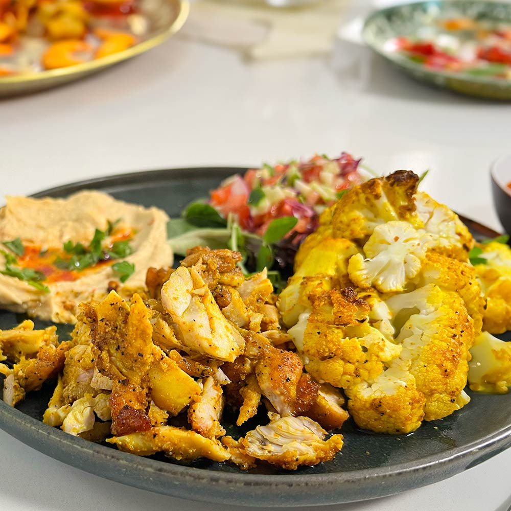 A plate of Mediterranean food with chicken shawarma and roasted cauliflower made with "Mama Shawarma" spice blend. Next to it hummus with paprika and garnish, and finely chopped salad.