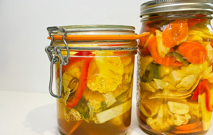 Two jars with pickled vegetables with the "In A Pickle" Mediterranean spice blend. The white cauliflower, garlic are yellowish from the healthy turmeric, along with pickled carrots, red pepper, and Jalapeños.