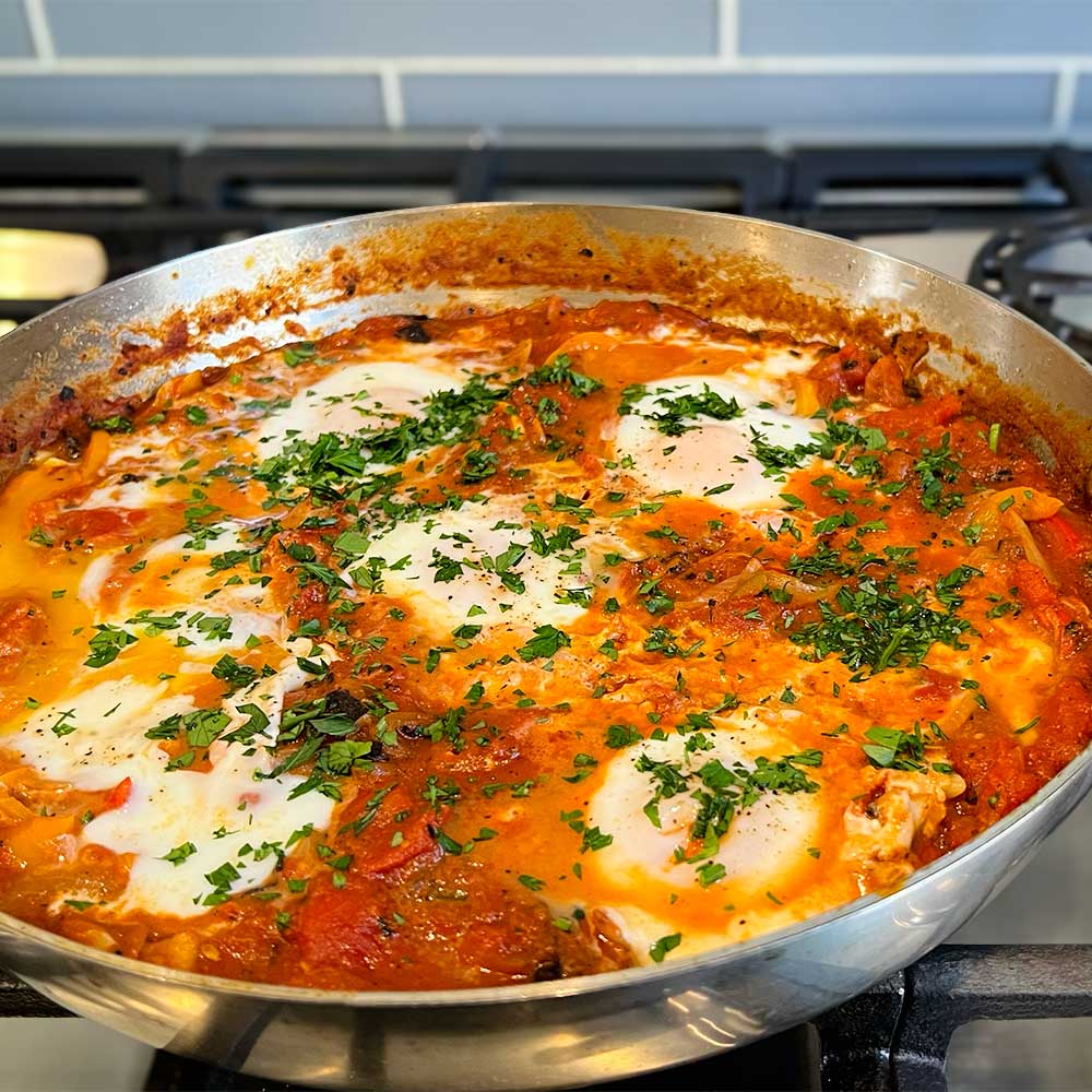 Stainless steel pan with red Mediterranean Shakshuka made with "Shake-Shuka" spice blend. Five poached eggs in the shakshuka sauce with chopped parsley garnish.